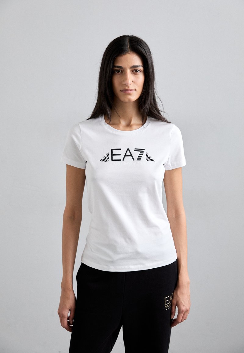 Balta trumpa rankovėmis marškinėliai su juodu "EA7" logotipu ir juostelėmis. Minkšta medvilnė, prigludęs dizainas, derinami su juodomis kelnėmis.