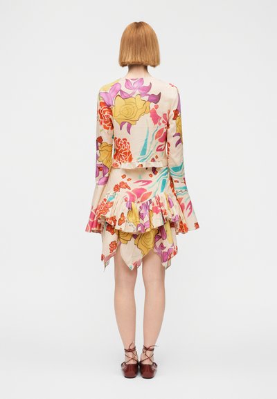 Γυναίκα με κοντά ίσια μαλλιά, φοράει φλοράλ μπλούζα με μακριά μανίκια και ασύμμετρη φούστα, στέκεται με την πλάτη προς την κάμερα σε λευκό φόντο.