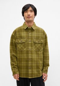 Chemise à carreaux verts en tissu doux, dotée de deux poches sur la poitrine, de fermetures à boutons et d'une coupe décontractée. Des coutures décoratives sont visibles partout.