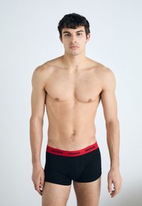 Schwarze Boxer-Briefs mit einem roten Markenbund. Das Modell steht mit freiem Oberkörper und entspannten Armen. Einfaches Design mit glatter Textur.