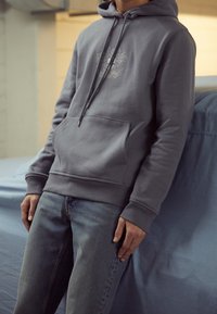 Felpa grigia con cappuccio, tasca frontale, cordoncino e polsini a coste, abbinata a jeans azzurri chiari. Design decorativo sottile sul petto.