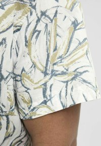 Camisa de manga corta de tela blanca con un patrón abstracto de hojas en azul y dorado, que presenta un corte holgado y un acabado texturizado.
