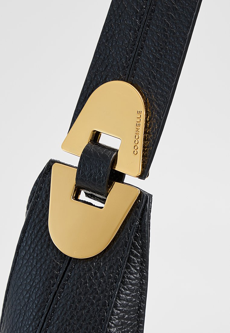 Cinghia della borsa in pelle nera con una grande fibbia in metallo dorato dal design distintivo. La superficie texturizzata aggiunge interesse visivo.