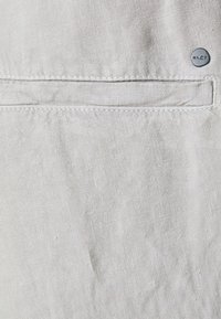 Jasnobeżowa tkanina z poziomą kieszenią i małym okrągłym metalowym guzikiem oznaczonym "NN07" w górnym prawym rogu.