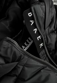 Juodas pūkinis švarkas su siuvinėta tekstūra viduje. Yra sidabrinė užtrauktukas ir prekės ženklo etiketė, ant kurios užrašyta "TED BAKER" balta spalva.