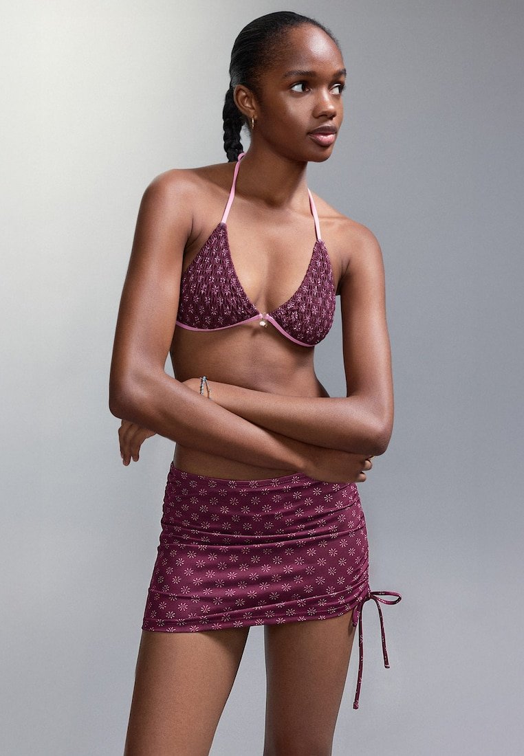 Femme aux cheveux tressés portant un haut de bikini à motifs bordeaux assorti à une mini-jupe froncée, debout avec les bras croisés, regardant vers la droite.