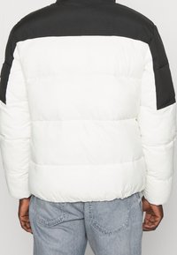 Osoba ubrana w czarno-białą kurtkę puchową i niebieskie dżinsy, pokazana od tyłu na jednolitym tle.