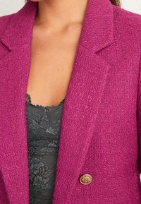 Veste en fuchsia texturée avec un col à revers et un détail de bouton doré. En dessous, un haut en dentelle noire avec des bords festonnés est visible.