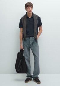 Herroutfit med en mörk polo, grå jeans och mörka mockaskor. En grå tröja är draperad över axlarna. En svart toteväska hålls.