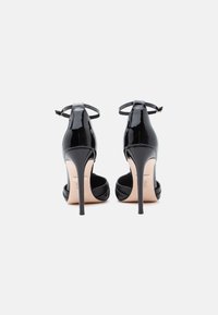 Must lineeritud mustad lakknahast kõrge kontsaga sandaalid, millel on pahkluuringid, stilletto kontsad ja beežid tallad, nähtud tagantpoolt valgel taustal.