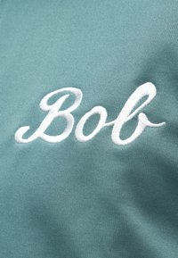 Λευκό κεντημένο κείμενο με καλλιγραφικά γράμματα "Bob" σε φόντο από υφασμάτινη υφή τυρκουάζ.