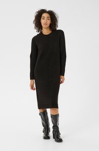 Robe pull noire longue et cintrée avec un col rond et des poignets côtelés. Portée avec des bottes noires montantes ornées de boucles.