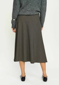 Jupe verte olive, longueur genoux, au design fluide et en tissu texturé. Associée à un pull gris, des chaussures plates noires visibles à l'ourlet.