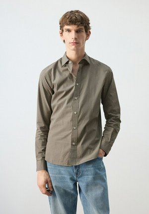 Joven con camisa marrón de cuadros de manga larga y jeans azul claro, de pie con una mano en el bolsillo contra un fondo liso.