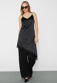 Vestido de satén negro con un dobladillo asimétrico fluido, decorado con encaje en la parte inferior. Combinado con pantalones negros de pierna ancha y zapatos negros.