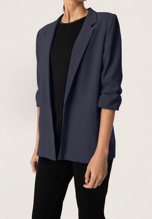 Femme portant un blazer bleu marine aux manches retroussées sur un haut noir côtelé et un pantalon noir, debout devant un fond clair uni.