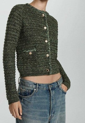 Personne portant un cardigan court en maille verte avec des boutons dorés et un jean bleu taille haute, main dans la poche, sur un fond uni.