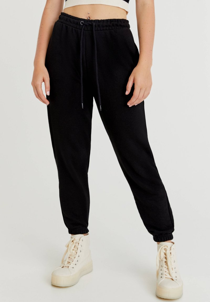 Personne portant un pantalon jogger noir avec cordon de serrage et poignets élastiques, et des baskets à plateforme montantes blanc cassé sur un fond uni.