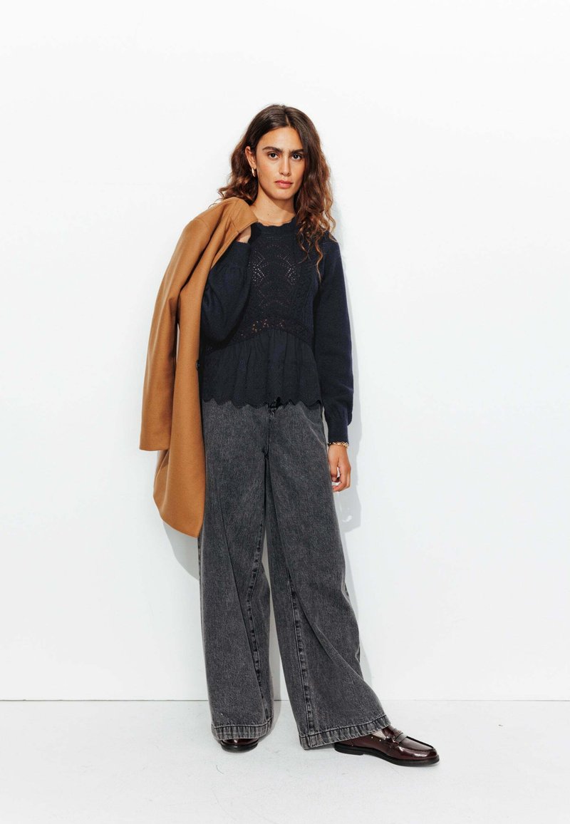 Haut en tricot texturé bleu marine superposé à un peplum avec un ourlet en dents de scie, associé à un pantalon en denim gris à jambes larges et un manteau marron drapé sur l'épaule.
