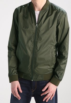 Man wearing zipped olive green bomber jacket over white shirt and dark blue jeans, standing against a plain background.