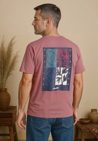 Roze katoenen T-shirt met een grafisch ontwerp op de achterkant met tropische patronen in blauw, rood en wit. Korte mouwen, casual pasvorm.