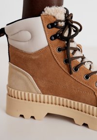 Bottine en nubuck marron avec des accents beiges, col en texure douce et duveteux, semelle en caoutchouc durable et lacets ronds. Détails cousus visibles.