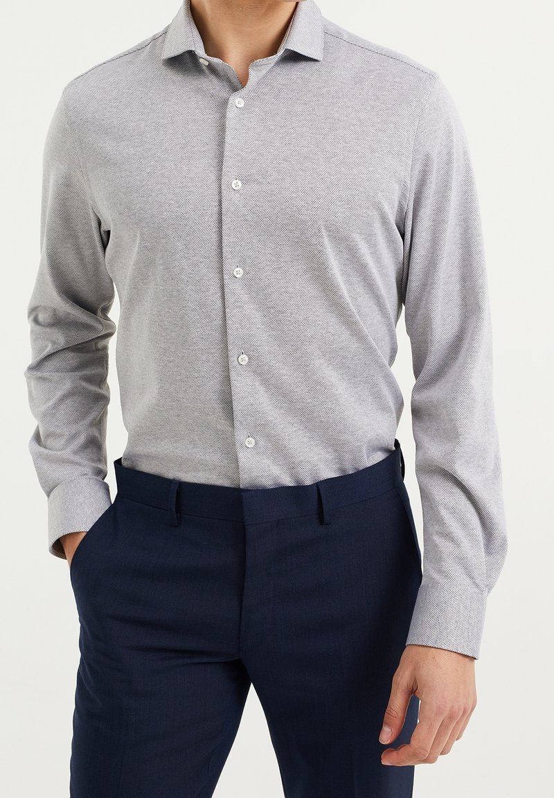 Man med ljusgrå strukturerad knapp-up skjorta instoppad i mörknavy byxor, med en hand i fickan mot vit bakgrund.