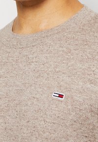 Beige stickad tröja med rund halsringning och en liten rektangulär röd, vit och blå logotyp på bröstet.