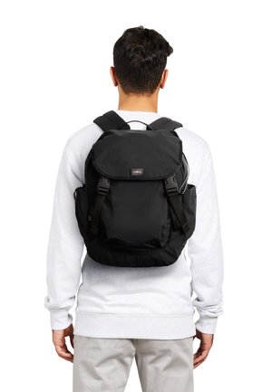 Uomo con felpa bianca e pantaloni beige con zaino nero dotato di patta superiore e tasche laterali, visto da dietro.