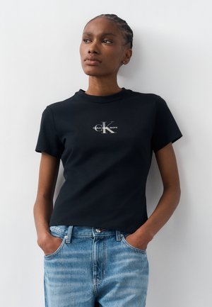 Νέα γυναίκα με πλεξούδες στα μαλλιά, φορώντας μια μαύρη μπλούζα Calvin Klein Jeans και ανοιχτόχρωμα μπλε τζιν, στέκεται με τα χέρια στις τσέπες.