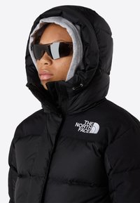 Persona con una chaqueta acolchada negra de The North Face con capucha, sudadera gris debajo y gafas de sol reflectantes negras, mirando hacia la izquierda.