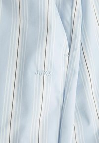 Svijetloplava majica s tkaninom s vertikalnim bijelim i crnim prugama. Sadrži izvezeni tekst "JJXX" u svijetloplavoj boji. Mekana tekstura, ležeran dizajn.