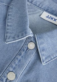 Šviesiai mėlynas džinsinis marškinėlių krūtinės apykaklė su matoma tekstūra, sidabrinėmis sagomis ir etiketė, ant kurios užrašyta "JJXX". Turi klasiką.