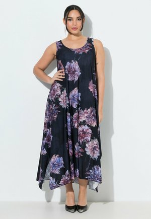 Frau steht mit einer Hand in der Hüfte, trägt ein ärmelloses schwarzes Maxikleid mit großem lila Blumenmuster und schwarze spitze Schuhe.