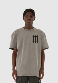 Grå oversized t-skjorte med rund hals, korte ermer, og en svart stilisert "m"-logo på venstre bryst. Lette teksturerte stoffer.