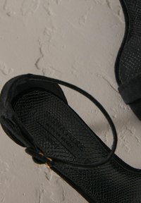 Sandalias de tacón alto negras con un acabado texturizado, que presentan una correa alrededor del tobillo y un sutil patrón tejido en la suela.