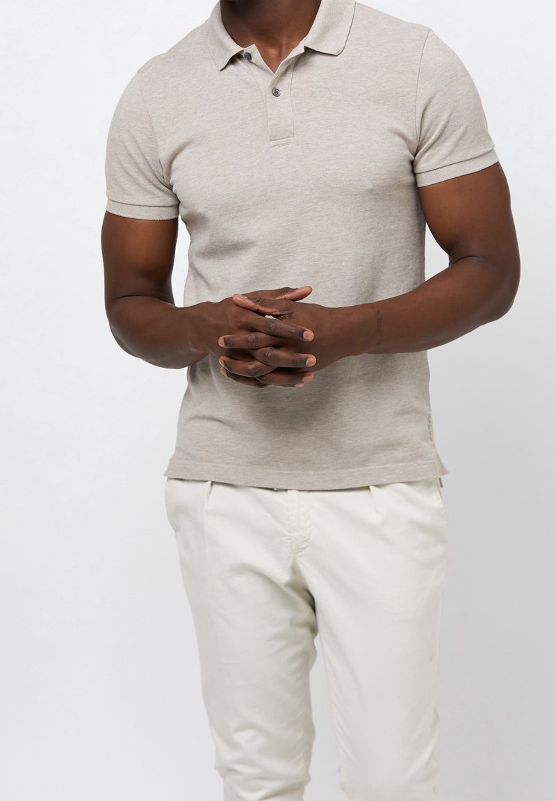 Homme portant un polo beige à manches courtes et un pantalon blanc, debout avec les mains jointes devant un fond uni.