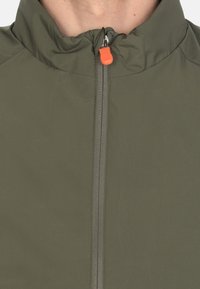 Giubbotto leggero verde oliva con colletto alto, zip frontale e un tirazip arancione. Tessuto liscio con dettagli minimi.