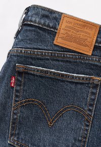 Jeans de ganga azul escuro com um remendo de cinto em pele castanha, apresentando costuras contrastantes em laranja e uma etiqueta vermelha "Levi's" no bolso traseiro.
