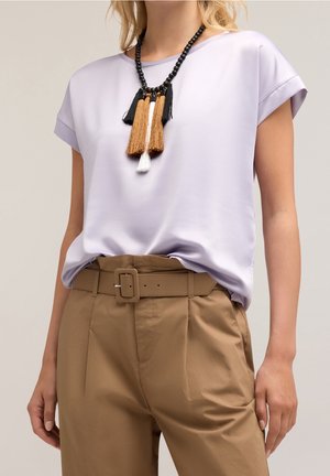 Femme portant un chemisier violet clair à manches courtes, un pantalon beige taille haute avec ceinture et un collier noir avec des pampilles marron, blanches et noires.