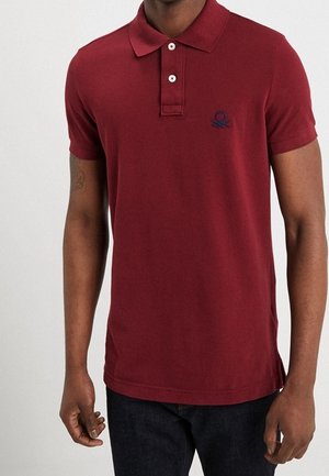 Mann trägt ein dunkelrotes Poloshirt mit kurzen Ärmeln, zwei Knöpfen und einem kleinen dunklen Logo auf der linken Brust.