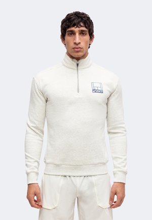 Homme portant un pull beige clair à demi-fermeture éclair avec le logo ASICS sur la poitrine et un pantalon beige clair assorti sur un fond uni.