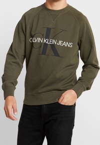 Man wearing olive green Calvin Klein Jeans sweatshirt with logo across chest and black jeans, hand in pocket.