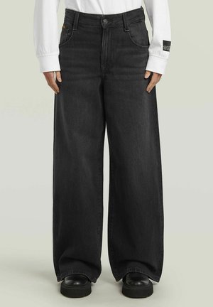 Persona che indossa jeans neri a gamba larga, scarpe nere massicce e una camicia bianca a maniche lunghe con una piccola toppa nera sulla manica sinistra.