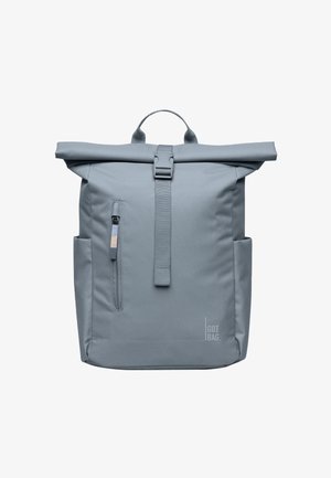 Sac à dos gris à rabat roulé avec poche zippée à l'avant, poches latérales, poignée supérieure, sangle centrale à boucle et marquage "GOT BAG" en bas à droite.