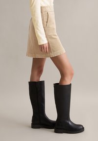 Zwarte kniehoge laarzen met een grove zool en een rits aan de zijkant, gedragen met een beige corduroy rok met een rechte pasvorm en zijzakken.