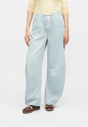 Person iført lyseblå brede bukser med striber, elastisk talje og snor, kombineret med brune sneakers, stående på hvid baggrund.