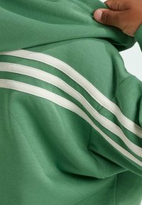 Zelená mikina s kapucí s třemi bílými pruhy na rameni. Materiál je hladký s mírně texturovaným povrchem a volným střihem.