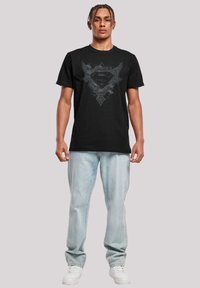 Black t-shirt featuring a grey graphic design of a shield with wings. Crew neck, short sleeves, paired with light blue jeans and white sneakers.