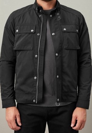 Mężczyzna w czarnej kurtce z przednimi kieszeniami z patkami, srebrnymi guzikami i szarą koszulą pod spodem, stoi z rękami luźno opuszczonymi wzdłuż boków.